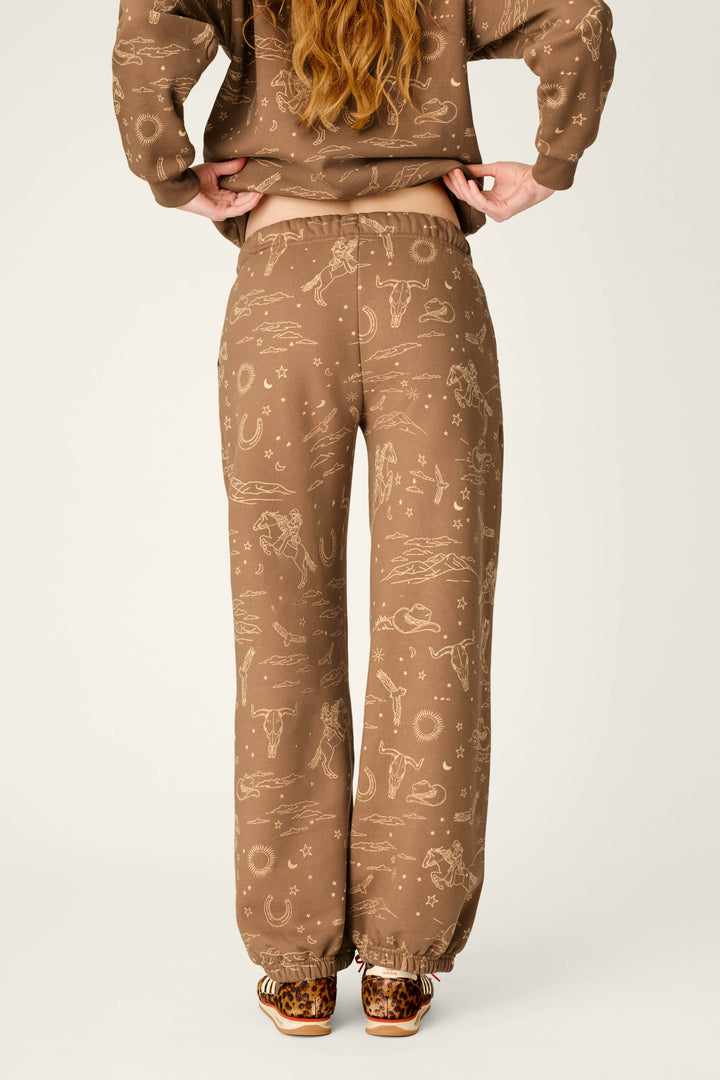 A person stands with hands on hips, wearing the P.J. Salvage Western Ways Ride ’Em Cowgirl Good Hang Joggers featuring beige western-style prints and a matching top. The loose-fitting pants are paired with leopard print slippers, and the image is taken from the back.