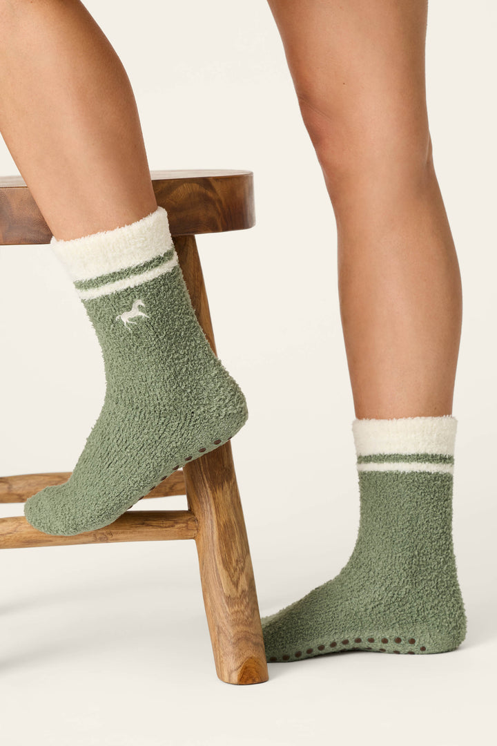 Two legs wear P.J. Salvage Wild Mustang Army Green Socks, featuring a fuzzy green fabric with white cuffs and stripes. One foot rests on a wooden stool while the other stands on the ground. The socks display a small animal logo and have non-slip grips on the sole.