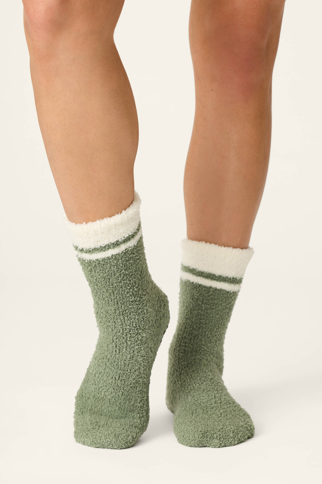 A person wearing P.J. Salvage Wild Mustang Army Green Socks, featuring soft, fuzzy fabric with white and green stripes at the top, standing on a light background.