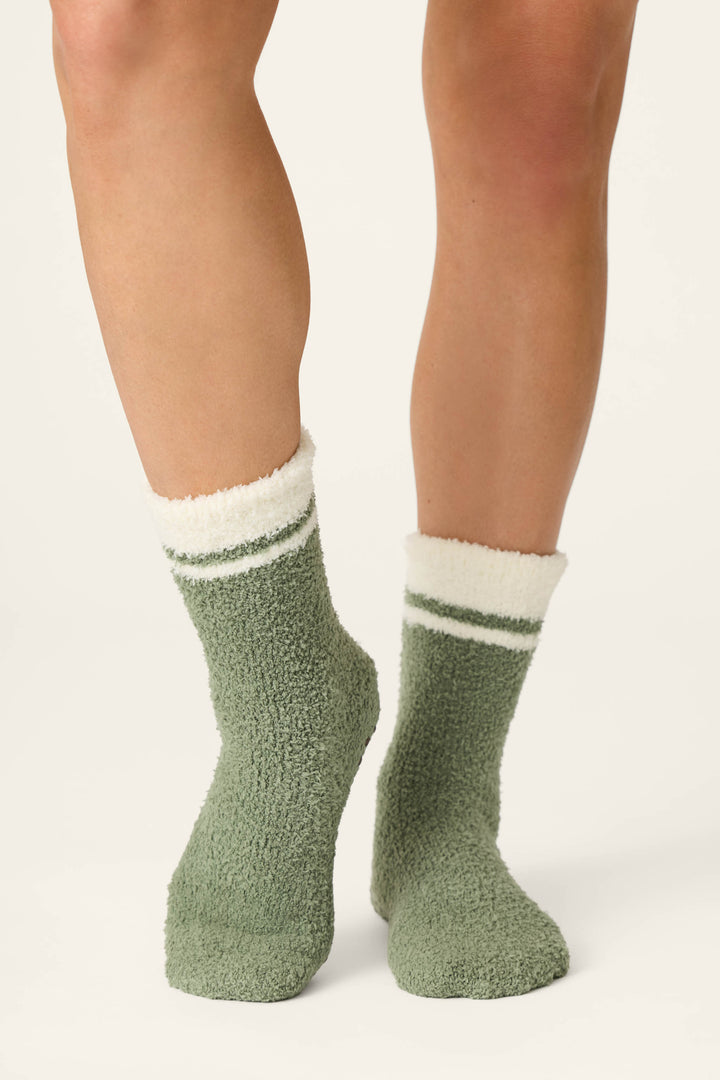 A person wearing P.J. Salvage Wild Mustang Army Green Socks, featuring soft, fuzzy fabric with white and green stripes at the top, standing on a light background.