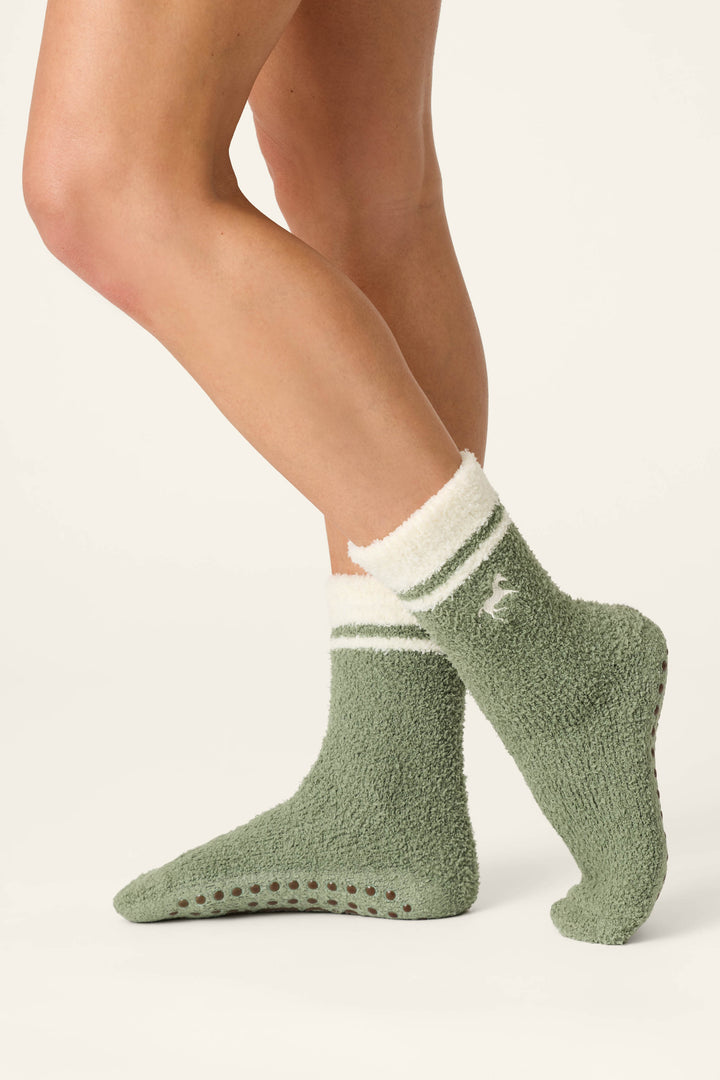 A person wearing P.J. Salvage Wild Mustang Army Green Socks, featuring white trim and non-slip grips on the soles, stands with one leg slightly bent in front of the other against a white background.
