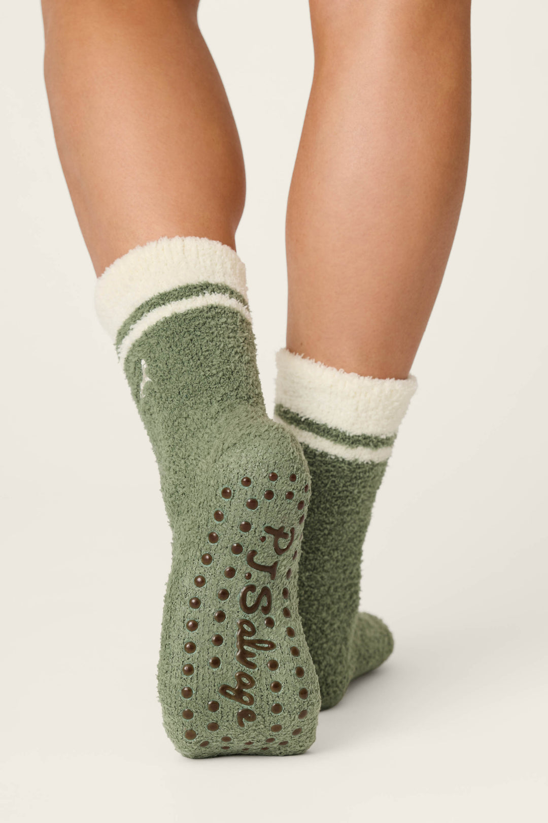 A person stands on tiptoe wearing P.J. Salvage Wild Mustang Army Green Socks, which feature fuzzy green fabric with white cuffs and stripes, non-slip grips, and the brand name on the soles.