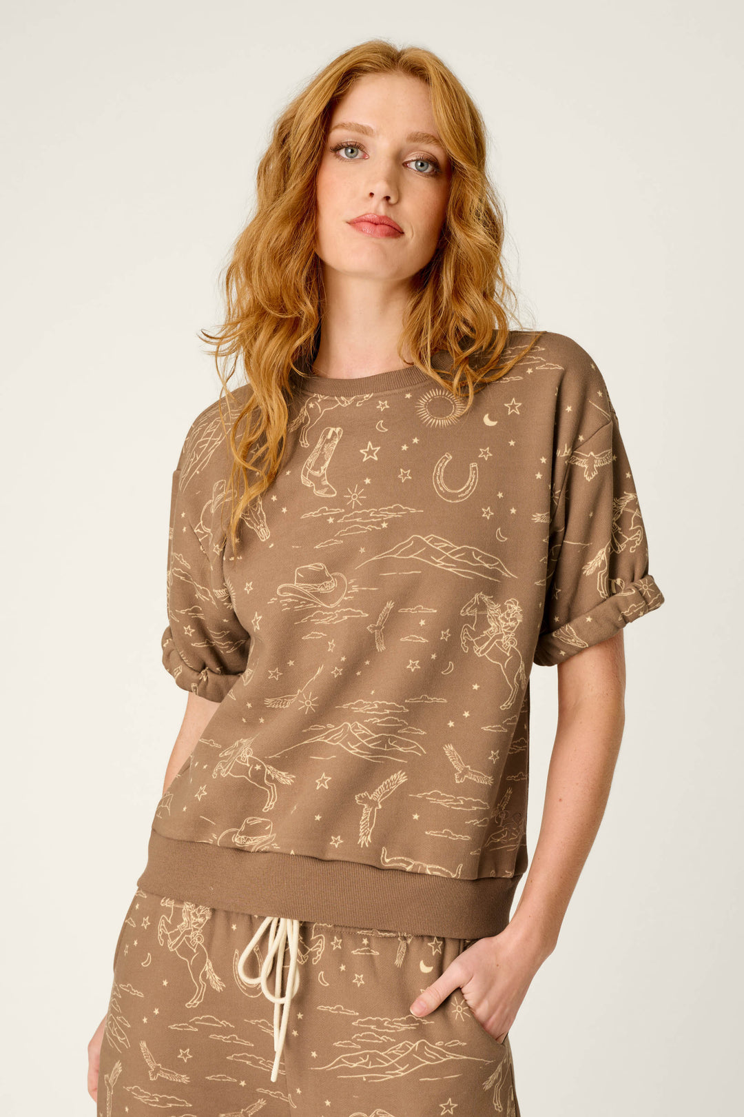 A woman with long red hair wears the P.J. Salvage Western Ways Ride ’Em Cowgirl Roll Back Short Sleeve Sweatshirt and matching pants, featuring beige Western-themed line drawings, while standing against a plain light background.
