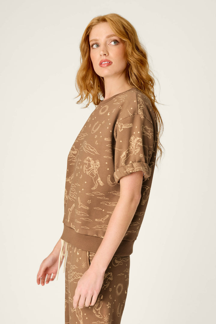 A woman with long red hair stands sideways and looks over her shoulder, wearing the P.J. Salvage Western Ways Ride ’Em Cowgirl Roll Back Short Sleeve Sweatshirt and matching pants adorned with light-colored celestial and animal patterns.