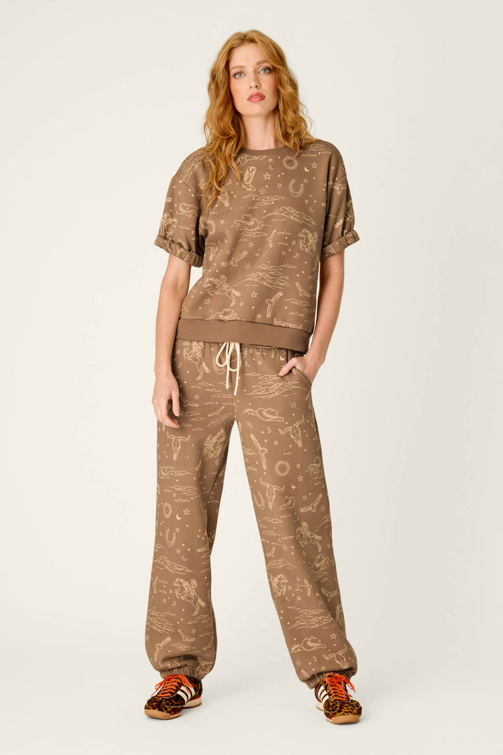 A woman with long red hair wears the P.J. Salvage Western Ways Ride ’Em Cowgirl Roll Back Short Sleeve Sweatshirt, styled with loose drawstring pants featuring light tan abstract patterns and orange animal print sneakers, standing against a plain light background.
