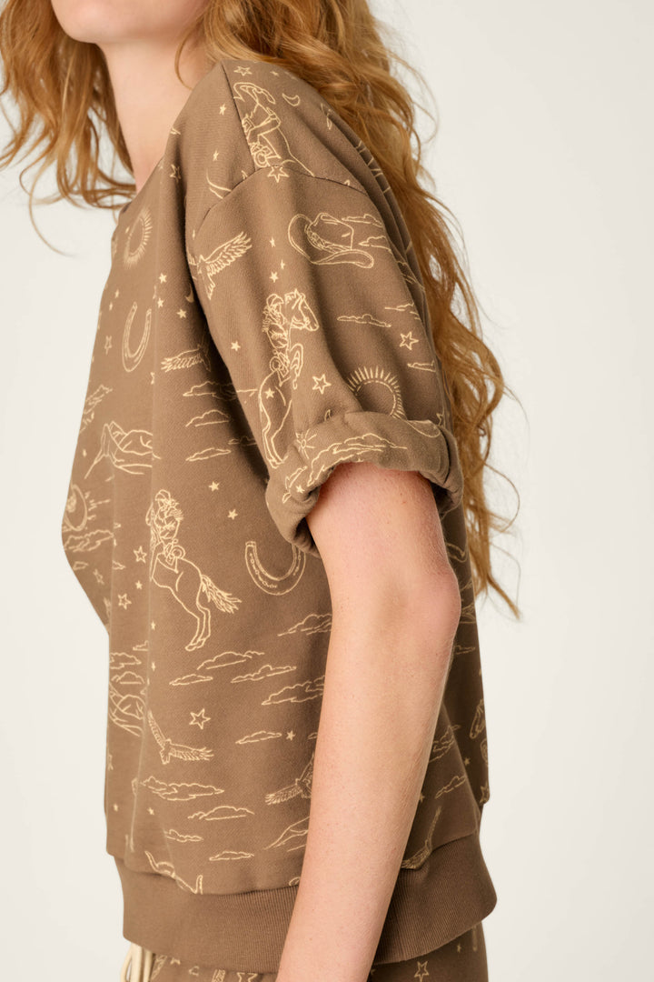 A person with long wavy hair wears the Western Ways Ride ’Em Cowgirl Roll Back Short Sleeve Sweatshirt by P.J. Salvage, a brown shirt adorned with a beige cowboy and western-themed print featuring horses, stars, and clouds. Only the torso and part of the face are visible.
