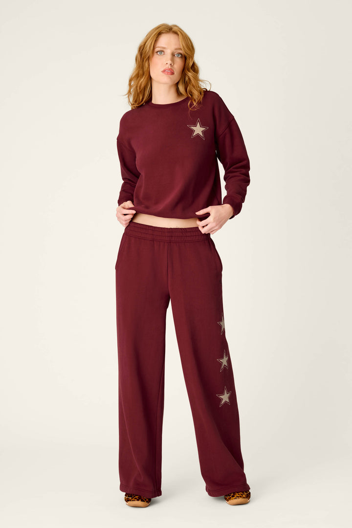 A woman poses in P.J. Salvage’s Western Ways Dark Wine BFF Lounge Pants, featuring star designs, paired with a matching maroon sweatshirt against a plain light background.