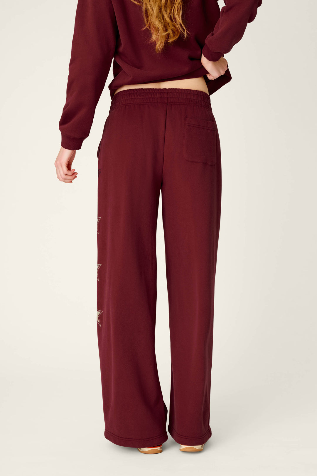 A person with long hair is shown from the back wearing the P.J. Salvage Western Ways Dark Wine BFF Lounge Pants, which feature a back pocket and star-shaped designs along the left leg, paired with a matching sweatshirt.