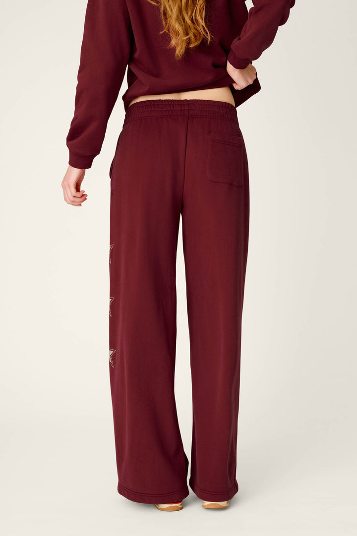 A person with long hair is shown from the back wearing the P.J. Salvage Western Ways Dark Wine BFF Lounge Pants, which feature a back pocket and star-shaped designs along the left leg, paired with a matching sweatshirt.