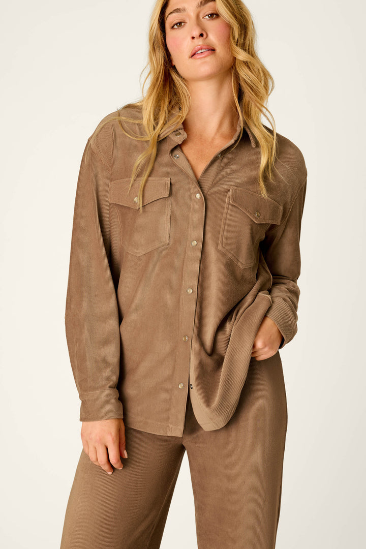 A woman with long blonde hair wears the P.J. Salvage Western Ways Hey Cowboy Corduroy Knit Button Down Top, featuring front pockets and a brown hue, along with matching brown pants, as she stands against a plain light background.