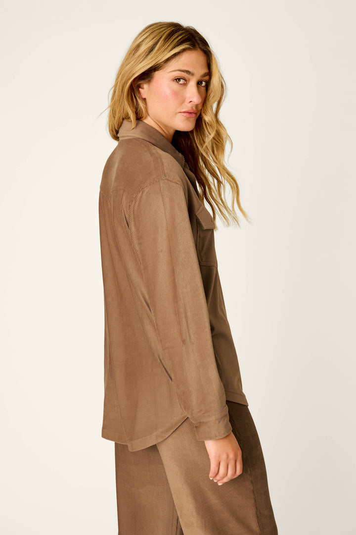 A woman with long, wavy blonde hair stands sideways against a plain light background, wearing the P.J. Salvage Western Ways Hey Cowboy Corduroy Knit Button Down Top in brown with matching pants and looking at the camera.