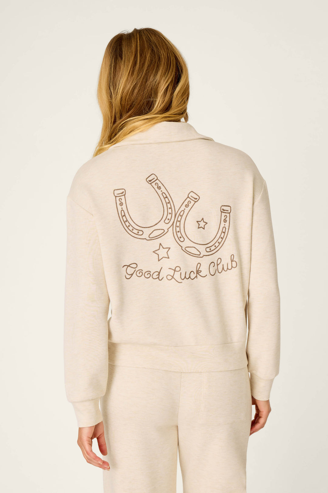 A woman with long blonde hair is shown from behind wearing the P.J. Salvage Western Ways Good Luck Club Over Night Pullover, a cream sweatshirt featuring two horseshoes, stars, and the words “Good Luck Club” printed on the back.