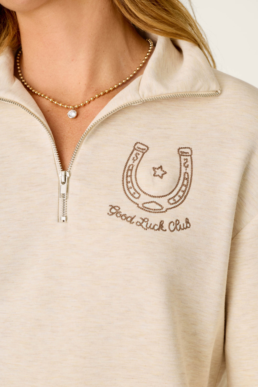 A person wearing the P.J. Salvage Western Ways Good Luck Club Over Night Pullover—a light beige zip-up sweatshirt with a horseshoe and “Good Luck Club” embroidery—paired with a gold beaded necklace featuring a small round pendant.
