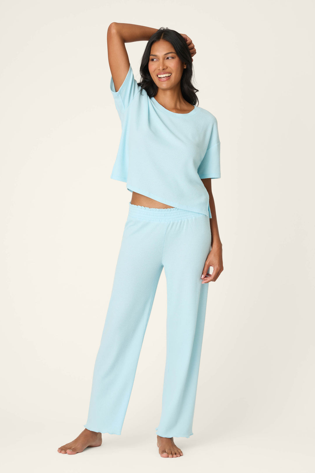 A woman stands barefoot on a light background, smiling with one arm raised. She wears P.J. Salvage’s Weekend Waffles Crystal Blue Pajama Pants, styled with a loose fit and wide legs.