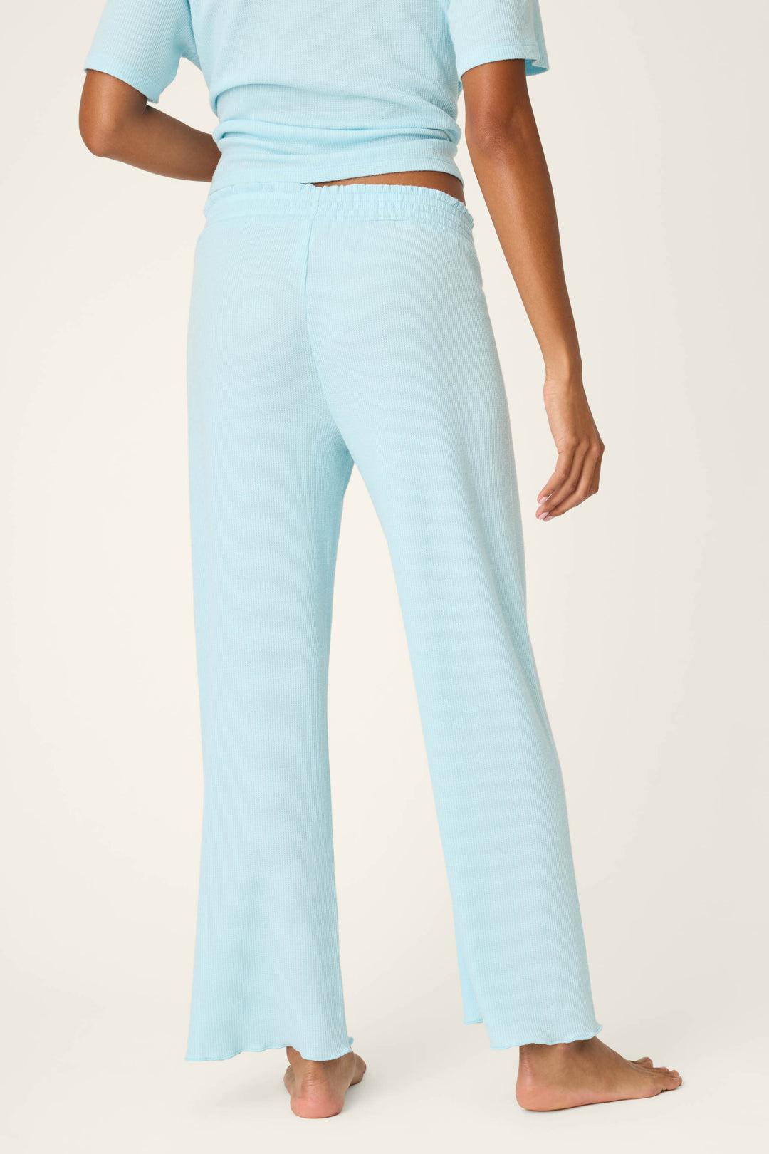 A person stands barefoot on a light surface, viewed from the back, wearing P.J. Salvage Weekend Waffles Crystal Blue Pajama Pants in a wide-leg fit with a matching short-sleeve top.