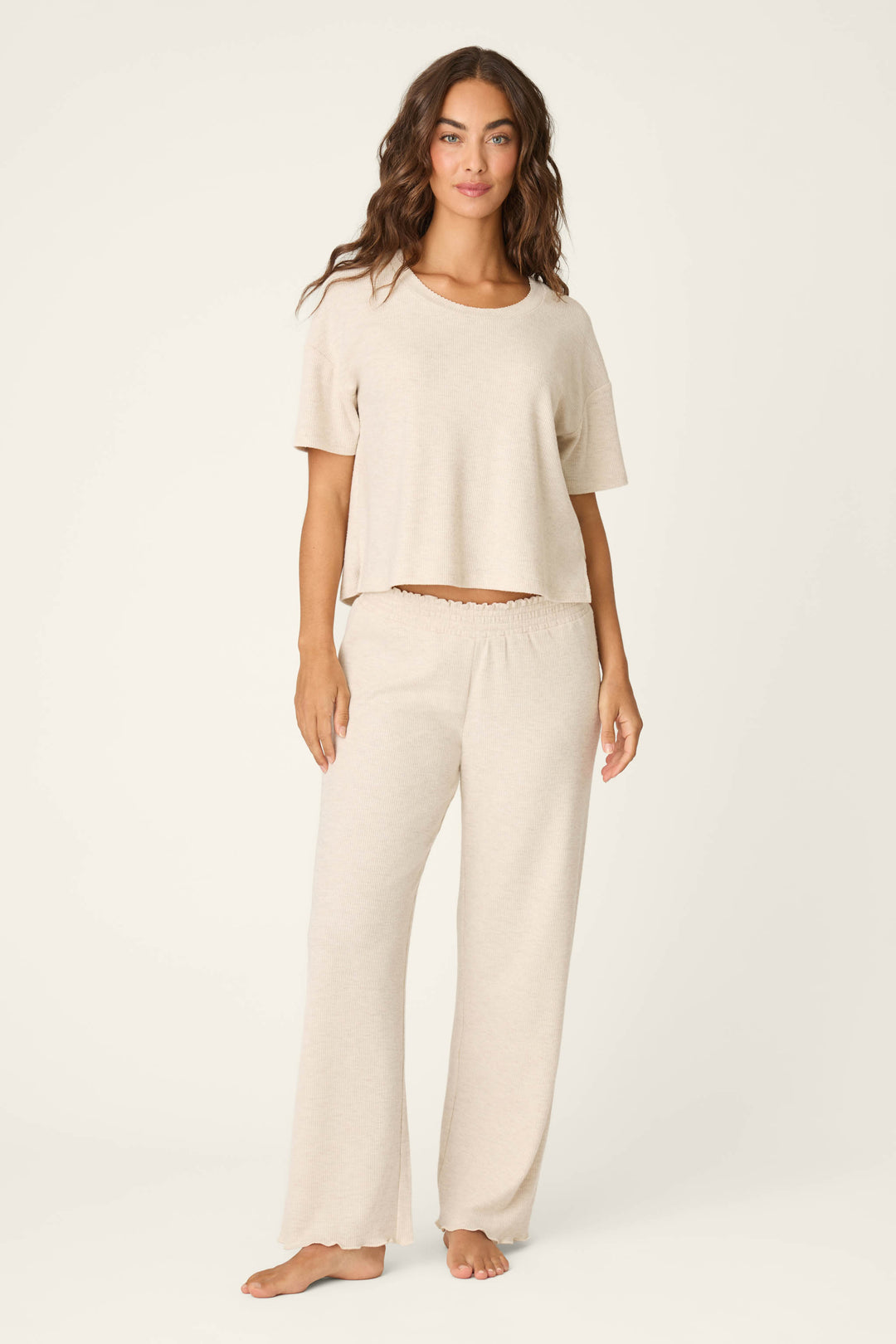 A woman stands barefoot against a plain background, wearing P.J. Salvage Weekend Waffles Light Oatmeal Pajama Pants with a loose fit, styled as loungewear and paired with a short-sleeved top.