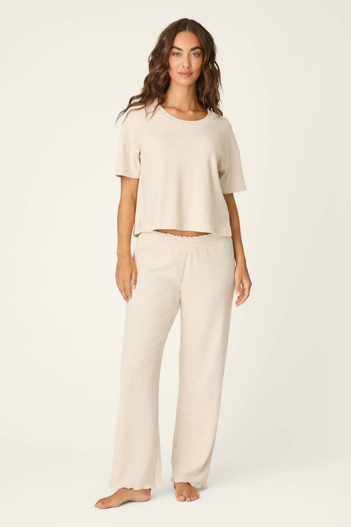 A woman stands barefoot against a plain background, wearing P.J. Salvage Weekend Waffles Light Oatmeal Pajama Pants with a loose fit, styled as loungewear and paired with a short-sleeved top.