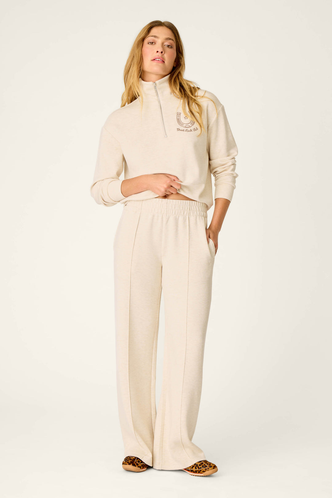 A woman stands in a relaxed pose with her hands in her pockets, wearing the P.J. Salvage Western Ways Good Luck Club Runway Lounge Pants in light beige paired with a matching half-zip sweatshirt. She has long, wavy hair and completes the look with leopard-print shoes against a plain, light-colored background.
