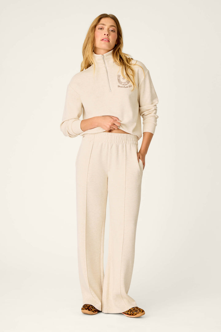 A woman stands in a relaxed pose with her hands in her pockets, wearing the P.J. Salvage Western Ways Good Luck Club Runway Lounge Pants in light beige paired with a matching half-zip sweatshirt. She has long, wavy hair and completes the look with leopard-print shoes against a plain, light-colored background.