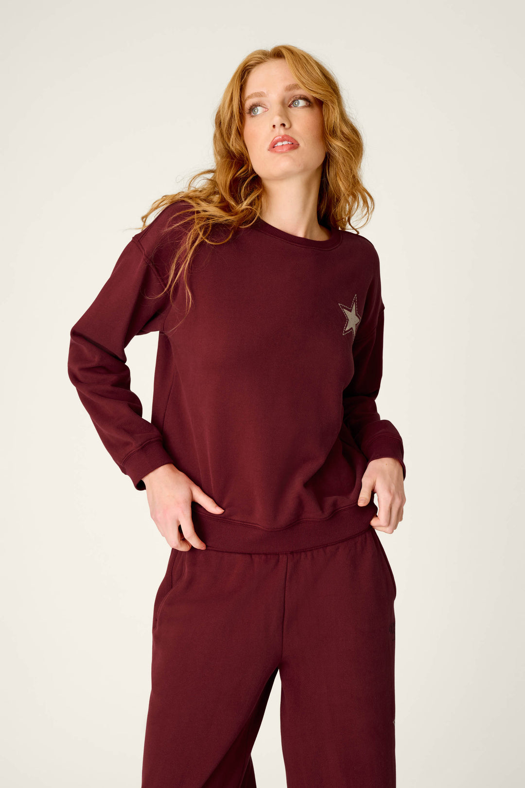A person with long, wavy hair wears the P.J. Salvage Western Ways Dark Wine Simone Sweatshirt, which features a small star patch on the chest, paired with matching sweatpants. They are looking slightly to the side against a plain, light-colored background.