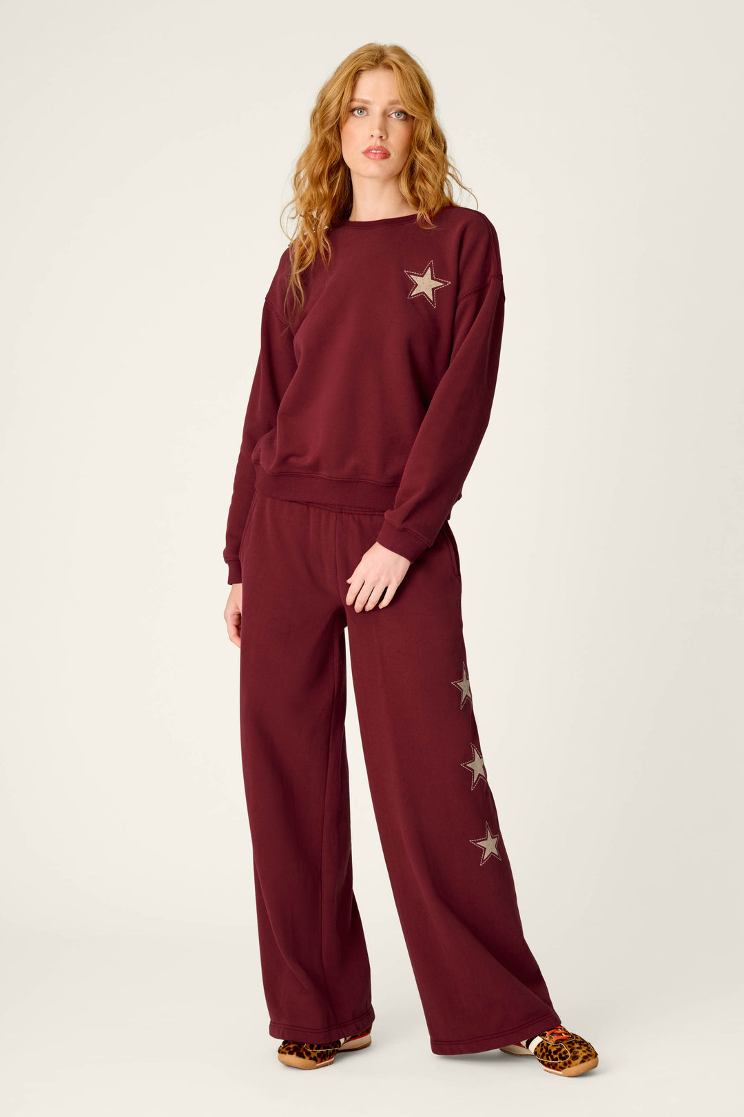 A woman with long wavy hair wears the Western Ways Dark Wine Simone Sweatshirt by P.J. Salvage, featuring star patches on the chest and leg, paired with leopard print slippers, standing against a plain light background.