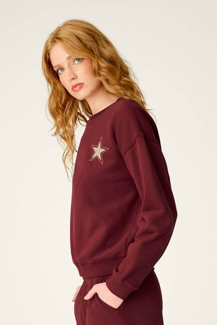 A woman with long wavy red hair stands sideways, looking toward the camera against a plain light background. She is wearing the P.J. Salvage Western Ways Dark Wine Simone Sweatshirt, which features a star patch on the chest, paired with matching pants.