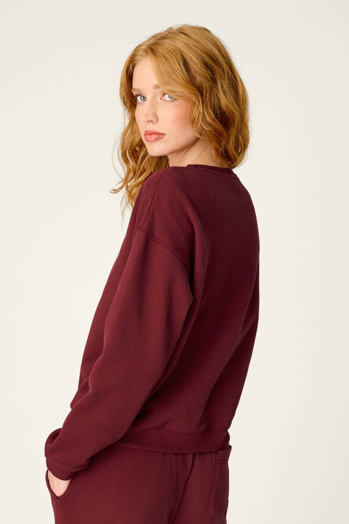Wearing the P.J. Salvage Western Ways Dark Wine Simone Sweatshirt, a person with wavy red hair and matching pants gazes over their shoulder against a plain light background.