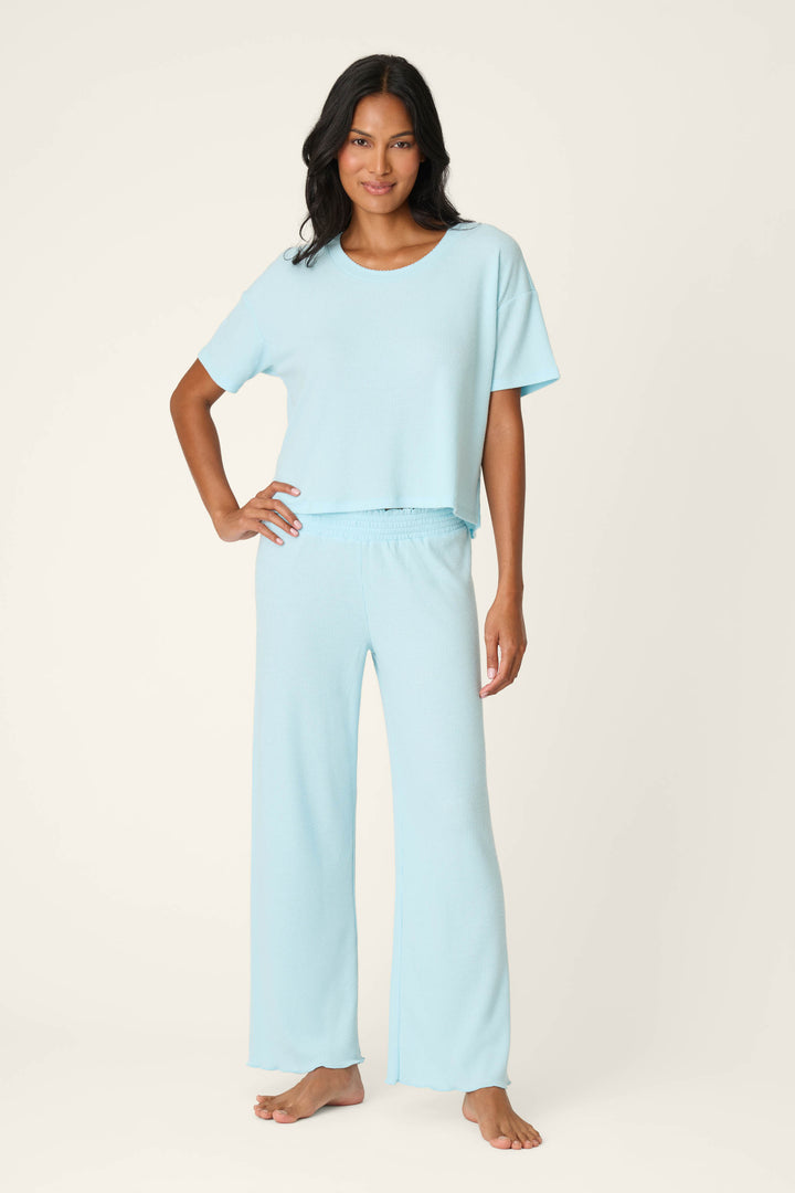 A woman stands barefoot against a light background, wearing the P.J. Salvage Weekend Waffles Crystal Blue Long PJ Set—a short-sleeve, loose-fit top and matching wide-leg pants—radiating relaxed loungewear vibes with a slight smile and one hand on her hip.
