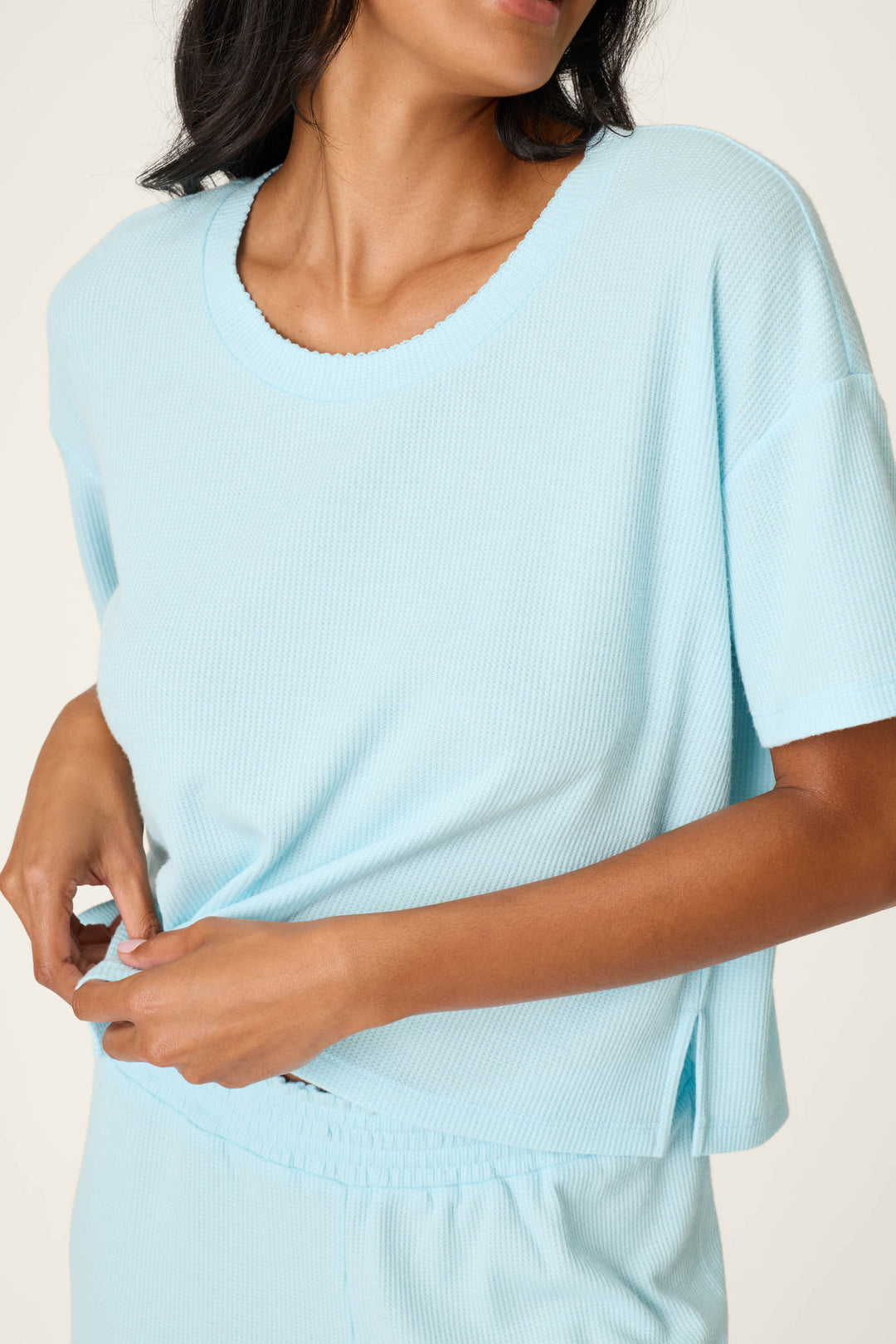A woman in the P.J. Salvage Weekend Waffles Crystal Blue Long PJ Set tugs at her top. The relaxed set includes a loose-fitting tee and matching elastic waist shorts, perfect for cozy lounging.