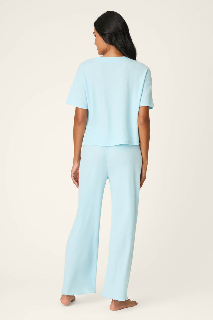 A woman with long dark hair stands barefoot, facing away, wearing the P.J. Salvage Weekend Waffles Crystal Blue Long PJ Set—short-sleeve top and matching wide-leg pants—against a plain beige background.