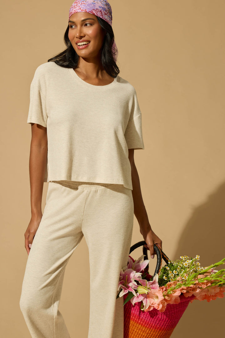 A woman in the P.J. Salvage Weekend Waffles Light Oatmeal Long PJ Set and a pink-patterned headscarf smiles while holding a colorful woven bag of flowers against a tan background—ideal for cozy, effortless style.