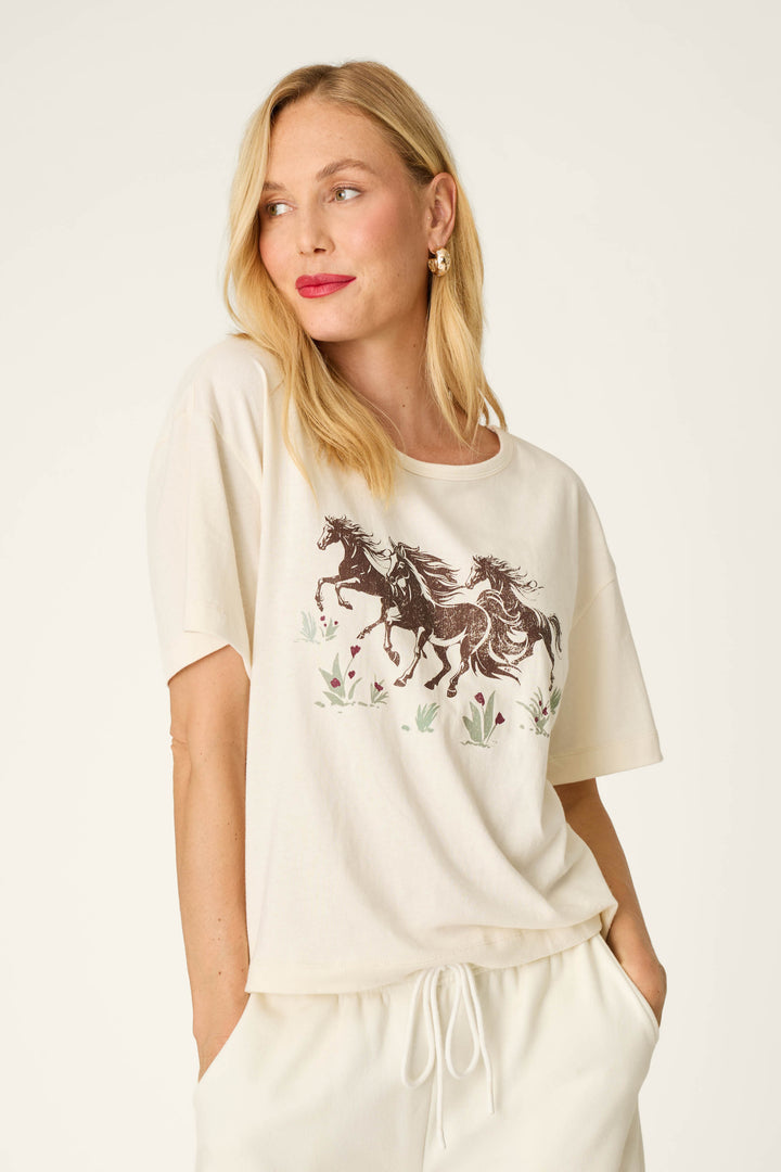 A woman with blonde hair is wearing the P.J. Salvage Western Ways Wild Horses Graphic Shuggie T-Shirt in cream, which features a print of two running horses and floral designs. She has paired it with matching light-colored pants, stands with her hands in her pockets, and gazes to the side.