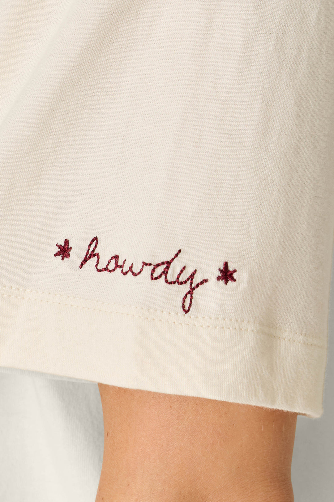 A close-up of the sleeve of the P.J. Salvage Western Ways Wild Horses Graphic Shuggie T-Shirt, featuring cream fabric and the word “howdy” embroidered in red cursive, accented by two small red stars.