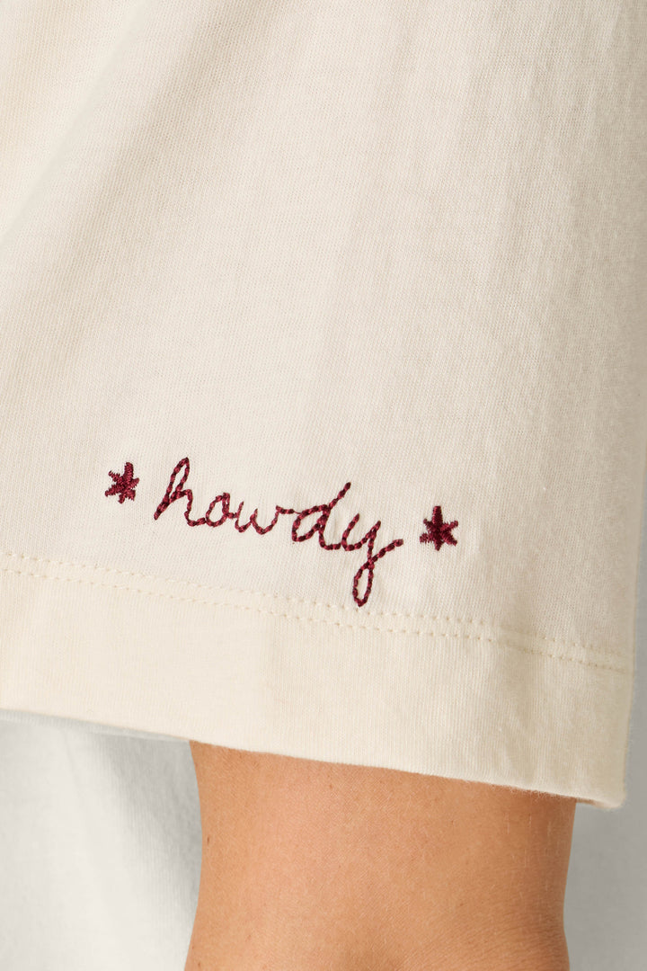 A close-up of the sleeve of the P.J. Salvage Western Ways Wild Horses Graphic Shuggie T-Shirt, featuring cream fabric and the word “howdy” embroidered in red cursive, accented by two small red stars.