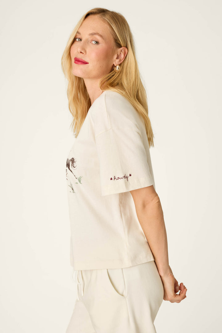 A woman with long blonde hair stands sideways and smiles softly against a plain background, wearing the P.J. Salvage Western Ways Wild Horses Graphic Shuggie T-Shirt in cream with matching pants.