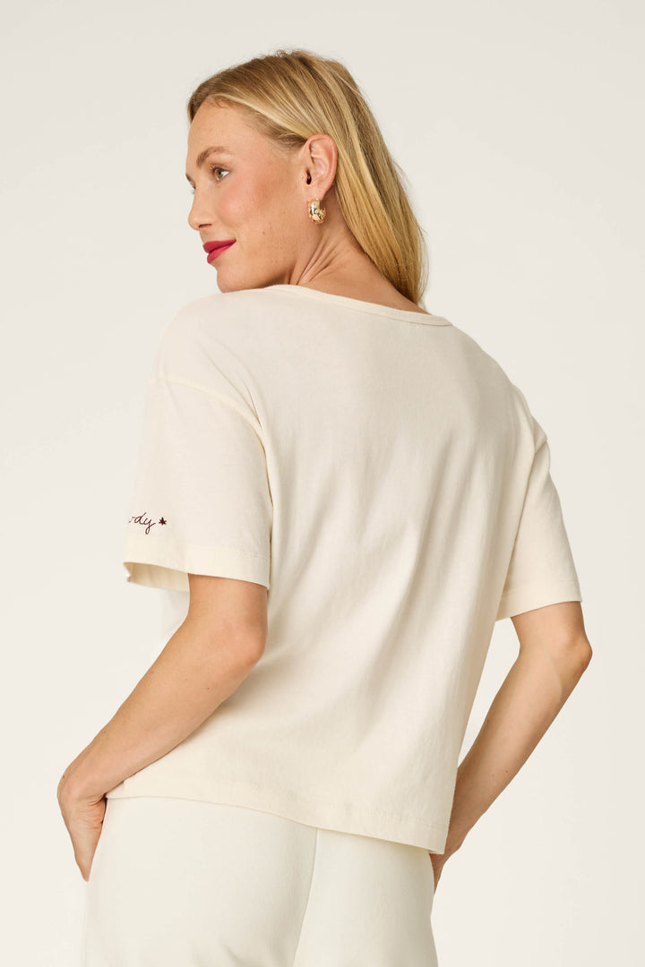 A woman with blonde hair is wearing the P.J. Salvage Western Ways Wild Horses Graphic Shuggie T-Shirt and matching pants, standing slightly turned away from the camera and looking over her shoulder against a plain light background.