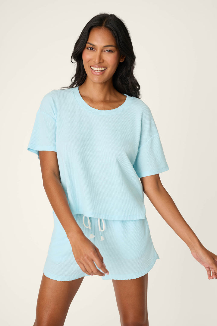 A woman smiles confidently in the P.J. Salvage Weekend Waffles Crystal Blue Short Sleeve Top, paired with matching drawstring shorts, against a simple light background.