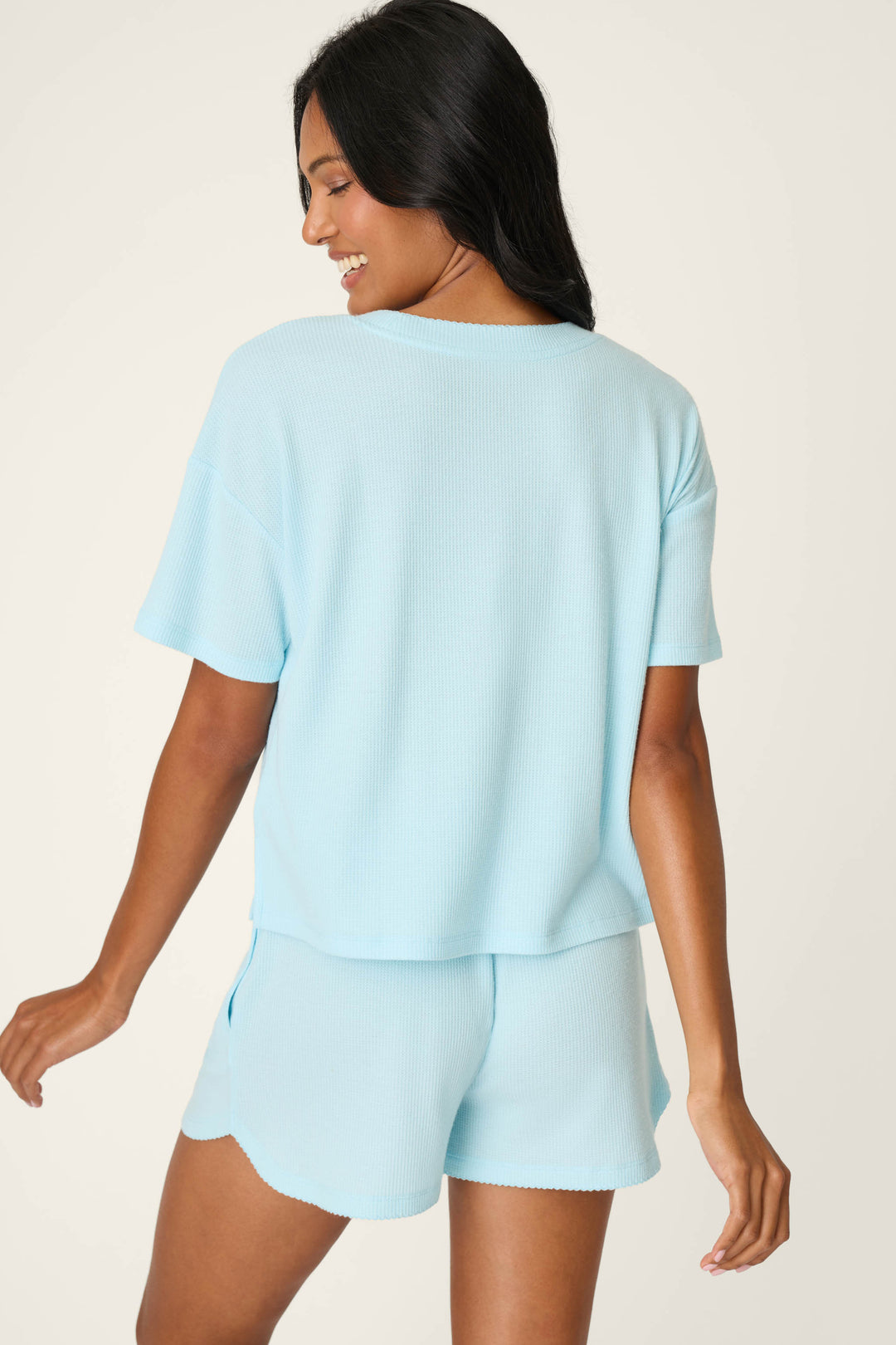 A woman with long dark hair wears the P.J. Salvage Weekend Waffles Crystal Blue Short Sleeve Top and matching shorts, standing against a light background and smiling over her shoulder.