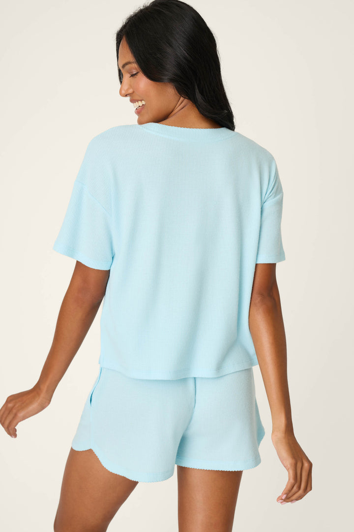 A woman with long dark hair wears the P.J. Salvage Weekend Waffles Crystal Blue Short Sleeve Top and matching shorts, standing against a light background and smiling over her shoulder.