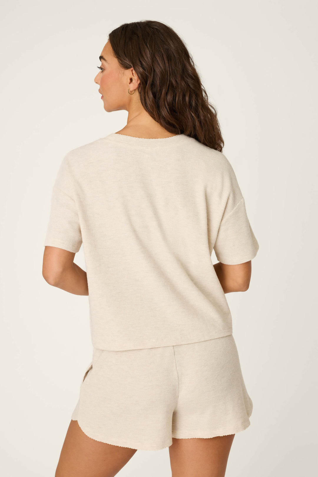 A woman with wavy brown hair stands with her back to the camera, wearing the P.J. Salvage Weekend Waffles Light Oatmeal Short Sleeve Top and matching shorts. The set features baby peachy waffle fabric, and the background is plain off-white.