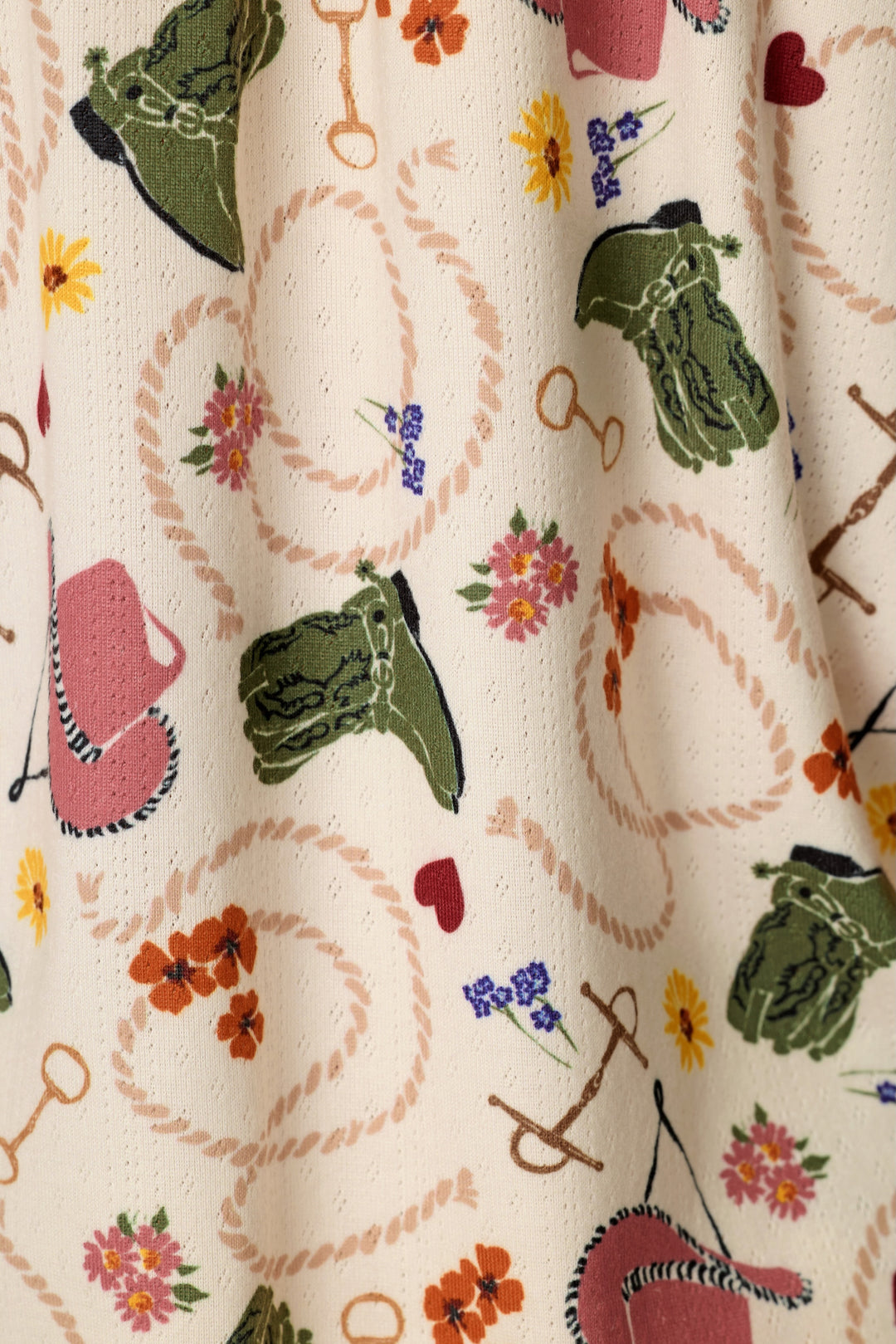 A close-up of a cream-colored fabric with a western-themed print featuring green and pink cowboy boots, rope loops, flowers, and small hearts.