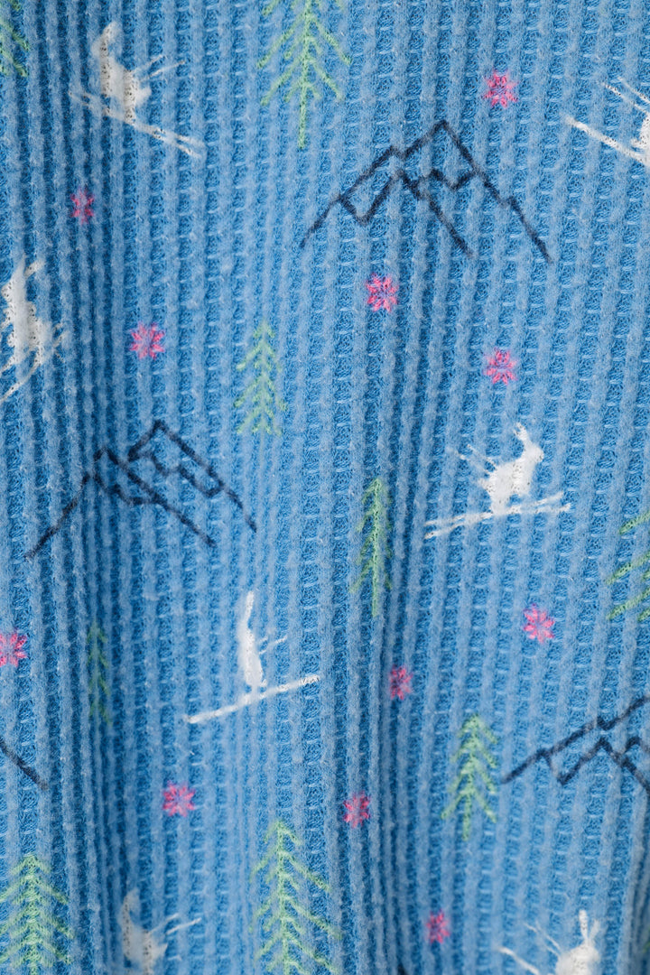 A close-up of a blue waffle-knit fabric with a winter sports pattern featuring skiing bunnies, pine trees, mountains, and pink snowflakes.