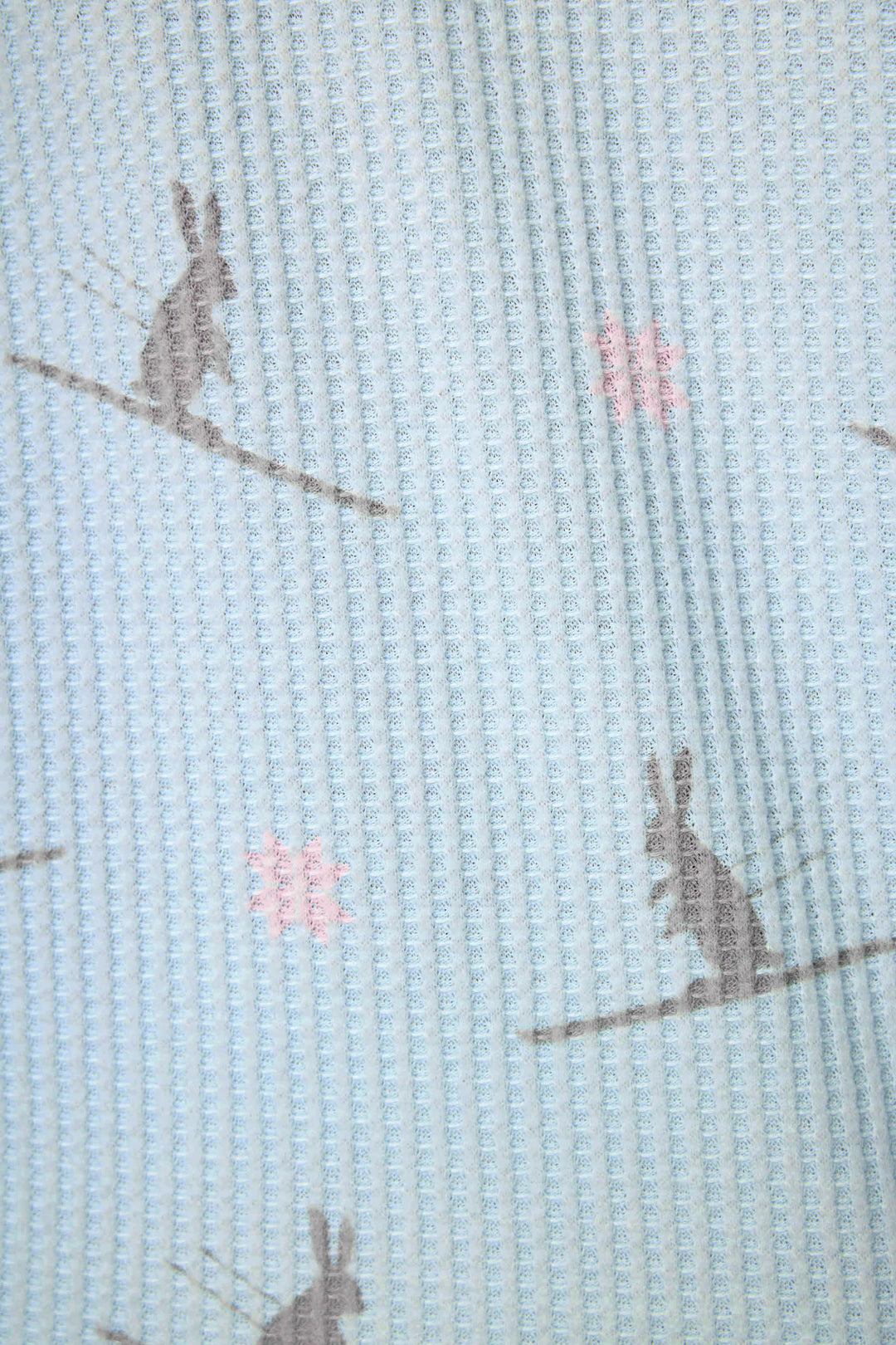 A close-up of the Ski Jammie Classics Ski Bunny PJ Set by P.J. Salvage, featuring light blue textured fabric with a repeating pattern of gray rabbits and small pink flowers. The simple design gives the pajamas a slightly wrinkled look.