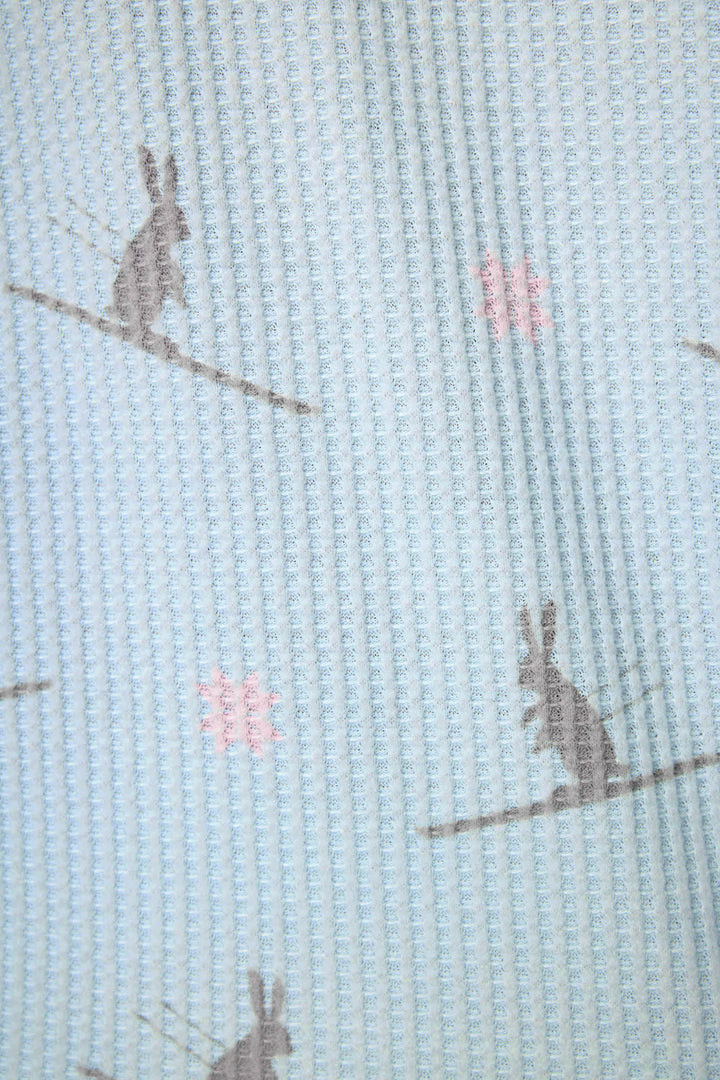 A close-up of the Ski Jammie Classics Ski Bunny PJ Set by P.J. Salvage, featuring light blue textured fabric with a repeating pattern of gray rabbits and small pink flowers. The simple design gives the pajamas a slightly wrinkled look.