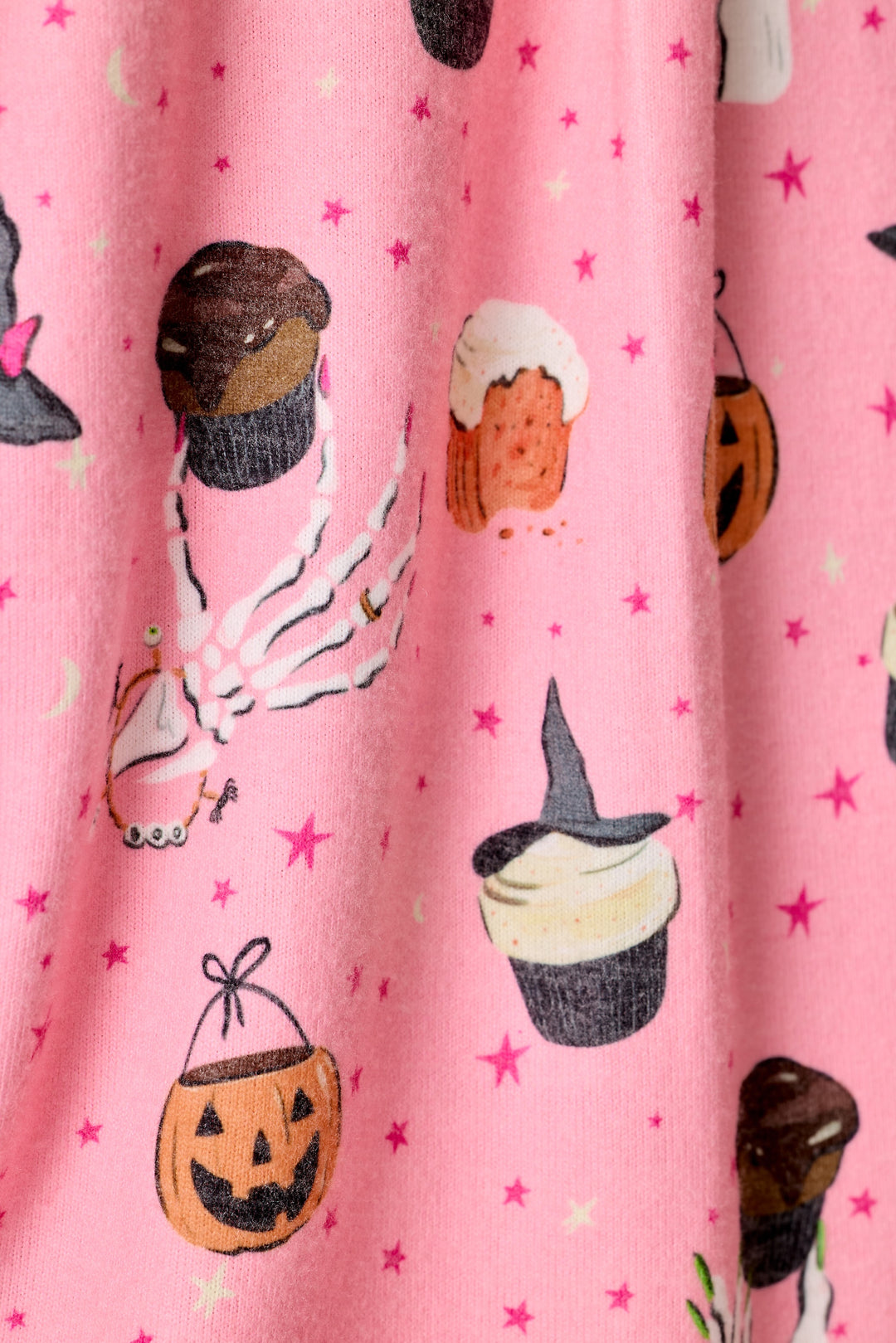 Close up of a pink fabric with a Halloween print featuring cupcakes, a jack-o'-lantern bucket, a witch hat, skeleton hands, and small stars.