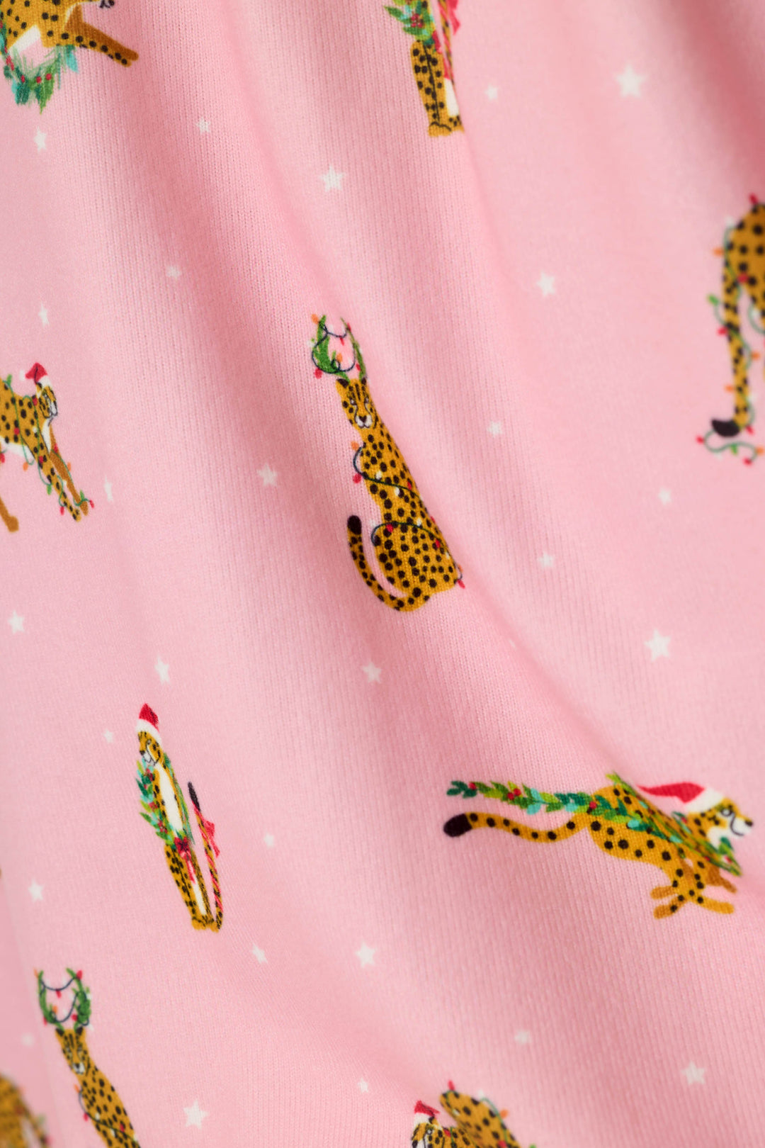 Pink fabric with a repeating print of yellow spotted cheetahs wearing green Christmas wreaths and red Santa hats, interspersed with small white stars.