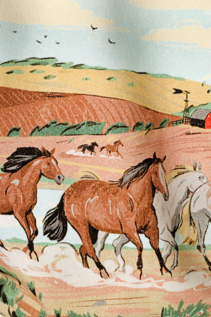 A close-up of a printed textile featuring a western landscape with several horses running through shallow water, hills, trees, birds, and a small red barn.