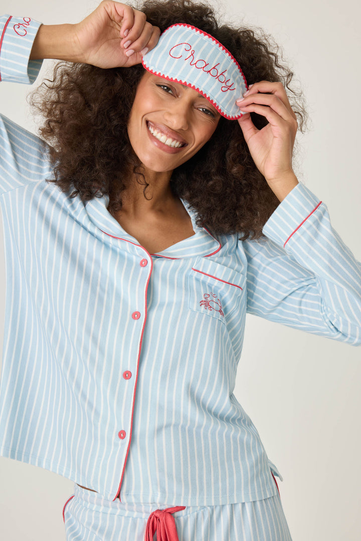Women's classic pajama set in light blue-white striped butter jersey. Red piping at button-front top & sideseams of pant. Chest pocket with embroidered crab & matching sleep mask with 'Crabby' lettering.