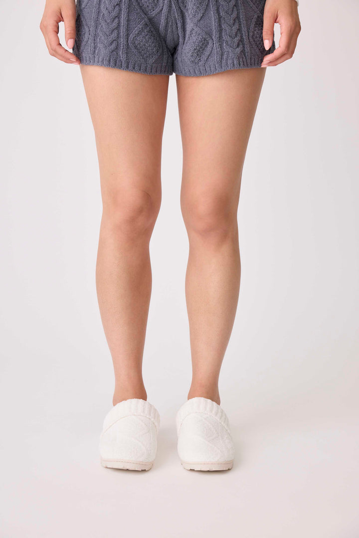 A person, visible only from the waist down, stands on a light background wearing blue knit shorts and P.J. Salvage Chenille Cozy Cable Slippers in white.